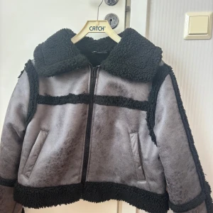 Grå teddyjacka med svarta detaljer - Säljer en grå teddyjacka med svarta, fluffiga kanter och foder. Jackan har dragkedja framtill, två fickor och en stor krage. Endast använd en gång. Passar mig som är en S/M. Tveka inte på att skriva prisförslag eller frågor!
