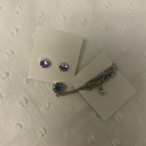 Lila örhängen och halsband med sten - Säljer ett set med silvriga örhängen och matchande halsband, båda med lila glittrande stenar. Perfekt för att ge en färgklick till din outfit. Kedjan är tunn och smidig med ett runt hänge.