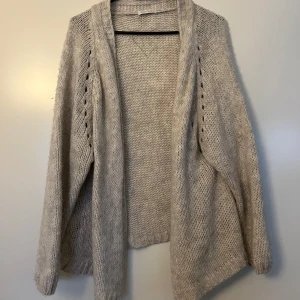 Cardigan, Heartkiss, grå, S/M - Ljusgrå tjock virkad cardigan från Heartkiss i storlek S/M. Fina öppna detaljer längs ärmarna. Avslappnad form och är öppen framtill. Mycket fint skick. 57% polyacryl, 29% polyamide, 7% viskos och 7% ull. Längd ca 65-75 (längre bak)