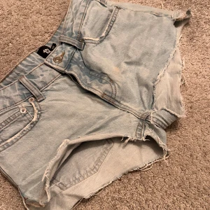 Jeansshorts - Säljer ett par ljusblå jeansshorts från 157 med råa fransade kanter och klassisk femficksmodell. Shortsen har låg midja och är perfekta för varma dagar. Snygg slitning och avslappnad look.