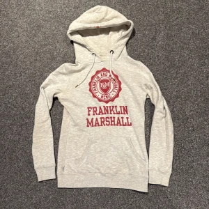 Franklin Marshall hoodie  - Grå hoddie med röd logga. Sitter skittajt på mig som man kan se på bild 2 och 3. Men fet hoodie för dig som har storlek S eller XS. Finns en fläck på bild 4.