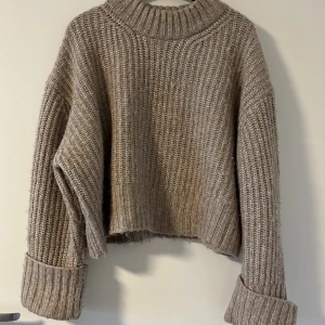Beige stickad croppad tröja - Säljer en beige stickad tröja med grov ribbstickning och croppad passform. Tröjan har breda uppvikta ärmslut och en hög, ribbad krage. Från Gina Tricot