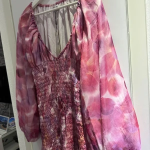 Rosa långklänning med mönster från NA-KD - Säljer en långklänning från NA-KD i rosa och lila toner med abstrakt mönster. Klänningen har långa puffärmar, smockad överdel och v-ringad hals. Perfekt för sommarens tillfällen!
