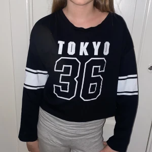 Svart croppad långärmad tröja med tryck från H&M - Säljer en svart croppad långärmad tröja från H&M med vitt tryck 'TOKYO 36' framtill och vita ränder på ärmarna. Perfekt för en sportig look och passar till både jeans och mjukisbyxor.