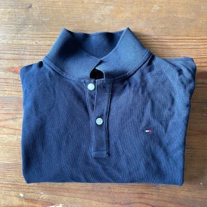 Mörkblå pikétröja från Tommy Hilfiger - Säljer en klassisk mörkblå pikétröja från Tommy Hilfiger med korta ärmar och två knappar framtill. Tröjan har en liten broderad logga på bröstet och detaljer i rött och vitt vid kragen.