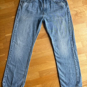 Levi's 505 ljusblå jeans - Klassiska ljusblå Levi's 505 jeans med rak passform. Jeansen har fem fickor, dragkedjegylf och tydliga slitningar för en vintagekänsla. Perfekta för dig som gillar en tidlös och avslappnad stil.