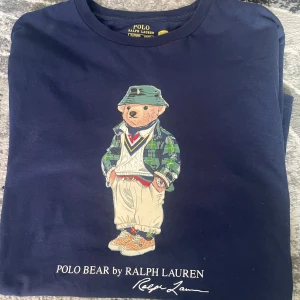 Mörkblå långärmad tröja med Polo Bear från Ralph Lauren - Säljer en mörkblå långärmad tröja från Ralph Lauren med Polo Bear-tryck framtill. Tröjan har rund halsringning och är i mjuk bomull. Klassisk och stilren design med ikonisk björn i streetwear-outfit.