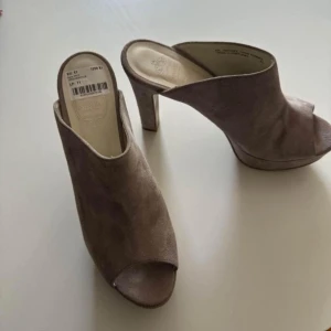 Beige mocka mules från Novita - Säljer ett par beige mules från Novita i mjuk mocka. Skorna har öppen tå, hög klack och en stilren design som passar perfekt till många outfits. Bekväma att bära och enkla att ta på tack vare slip-in modellen. Stl 41 men passade min fot som är 39 
