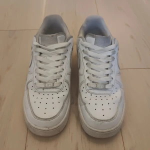 Vita Nike Air Force 1 sneakers  - Klassiska vita Nike Air Force 1 sneakers med rund tå och platt sula. Skorna har snörning och är tillverkade i skinn med perforerade detaljer framtill för extra ventilation. Perfekta för en stilren och sportig look.