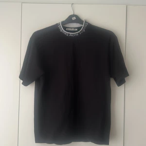 Svart t-shirt från Acne Studios med logga på kragen - Säljer en svart t-shirt från Acne Studios med deras logga broderad runt kragen. T-shirten har en klassisk passform och är tillverkad i mjuk bomull. Perfekt för dig som gillar stilrena in och trendiga plagg med diskreta detaljer.
