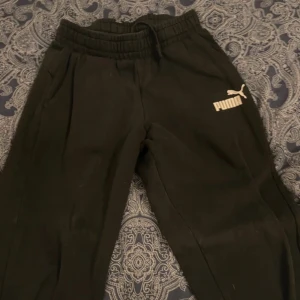 Svarta mjukisbyxor från Puma - Svarta mjukisbyxor från Puma med resår i midjan och dragsko. Byxorna har sidofickor och en vit Puma-logga på vänster lår. Perfekta för en avslappnad stil. Finns tre par två i storlek 14år och en i storlek xs