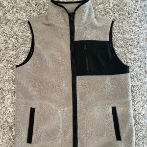 Beige väst  - Säljer en beige fleeceväst med svarta detaljer och dragkedja framtill. Västen har hög krage som går att vika ner, svarta fickor och en bröstficka med dragkedja. Perfekt för lager-på-lager under kyliga dagar.
