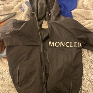 Svart vindjacka från Moncler - Säljer en svart vindjacka från Moncler med stor logotyp på bröstet och ärmen. Jackan har huva, dragkedja framtill och två fickor med dragkedja. Perfekt för dig som vill ha en stilren och exklusiv jacka.