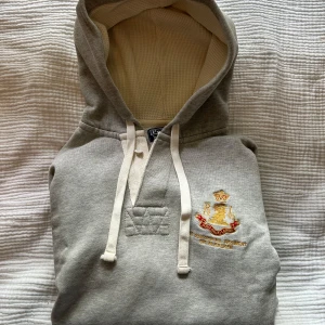 Grå hoodie från Ralph Lauren - Säljer en ljusgrå hoodie från Ralph Lauren med broderad logga och emblem i gult och rött på bröstet. Frugan har inga effekter. storlek M