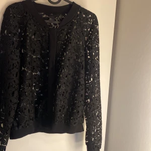 Svart spetskofta från H&M - Säljer en svart kofta från H&M i storlek M. Koftan är helspets med blommigt mönster och har ribbade muddar vid hals, ärmslut och nederkant. Perfekt för att lyfta en outfit med en cool och elegant touch.