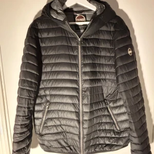 Svart pufferjacka från Colmar - Snygg svart pufferjacka från Colmar med huva och dragkedja framtill. Jackan har två fickor med dragkedja och ett diskret Colmar-märke på ärmen. Perfekt för kyliga dagar och enkel att matcha med olika outfits.