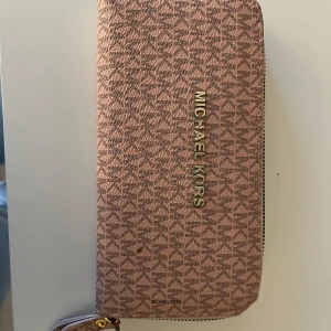 Rosa plånbok från Michael Kors - Säljer en snygg rosa plånbok från Michael Kors med guldiga detaljer och dragkedja runtom. Plånboken har MK-logga över hela ytan och är perfekt för att hålla ordning på kort och kontanter. Stilren och trendig accessoar!