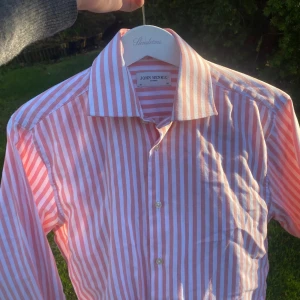 Mörk Rosa och vit randig skjorta från John Henric - Säljer en klassisk långärmad skjorta från John Henric med rosa och vita ränder. Skjortan har knappar framtill och en klassisk krage. Perfekt för dig som gillar stilrena och färgglada plagg.