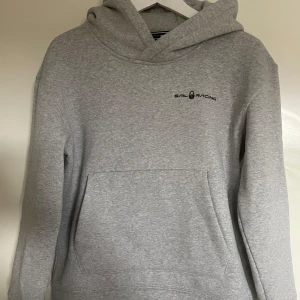 Grå hoodie från Sail Racing - Säljer en stilren grå hoodie från Sail Racing med svart logga på bröstet. Tröjan har huva och en stor magficka framtill. Perfekt för en avslappnad look och enkel att matcha med det mesta.