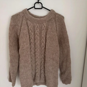 Beige stickad tröja med flätmönster - Mysig beige stickad tröja med flätmönster framtill och rund halsringning. Tröjan har långa ärmar och är perfekt för kyligare dagar. Passar till det mesta och är superbekväm. 