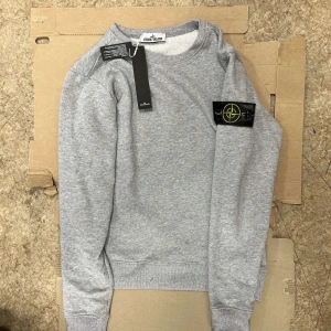 Stone island Sweater - Säljer en stilren grå sweatshirt från Stone Island med klassisk logotyp-patch på vänster ärm. Tröjan har rund halsringning och ribbade muddar vid ärmslut och nederkant. Perfekt för en avslappnad och trendig look.