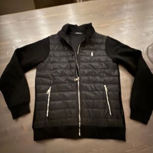 Svart quiltad jacka från Polo Ralph Lauren - Snygg svart jacka från Polo Ralph Lauren med quiltad front och ärmar i mjuk bomull. Jackan har dragkedja framtill, två fickor med dragkedja och en liten logga på bröstet. Perfekt för en stilren look.