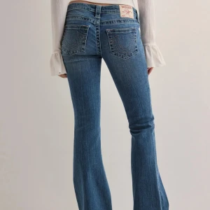 Blå bootcut jeans från True Religion - Säljer ett par klassiska blå bootcut jeans från True Religion med snygga kontrastsömmar och ikoniska bakfickor. Jeansen har låg midja och en figurnära passform upptill som går ut i ett bootcut nertill. Ändast använd 1 gång och är i jättebra skick. Ny pris 1399.