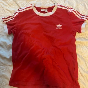 Adidas Originals T-shirt - Röd t-shirt från Adidas med vita ränder på axlarna och vita muddar vid hals och ärmslut. Klassisk Adidas-logga på bröstet. Perfekt för en sportig och avslappnad stil.