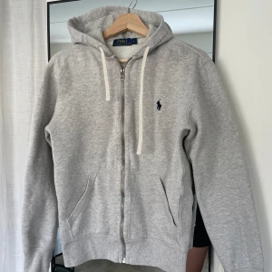 Grå hoodie från Polo Ralph Lauren - Säljer en klassisk grå hoodie från Polo Ralph Lauren med dragkedja och huva. Tröjan har två fickor framtill och en broderad marinblå logga på bröstet. Perfekt för en avslappnad stil.