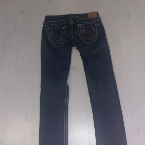 Jeans från True Religion - Säljer ett par mörkblå jeansbyxor från True Religion med kontrastsömmar och klassiska bakfickor med lock. Jeansen har raka ben och normal passform. Perfekta för dig som gillar en tidlös jeansstil.