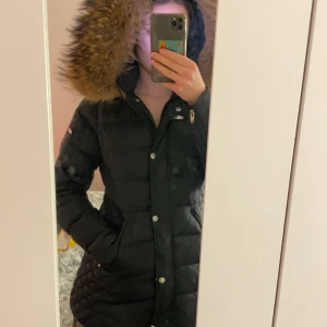 Svart dunjacka med pälskantad huva från Rockandblue - Säljer en svart dunjacka från Rockandblue med pälskantad huva och quiltade detaljer på sidorna. Jackan har dragkedja och knappar framtill samt fickor med dragkedja. Perfekt för kalla dagar! ÄKTA PÄLS!