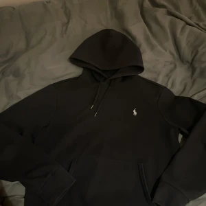 Svart hoodie från Polo Ralph Lauren - Säljer en svart hoodie från Polo Ralph Lauren med klassisk broderad logga på bröstet. Tröjan har huva med dragsko och en stor ficka framtill. Perfekt för en avslappnad och stilren look.