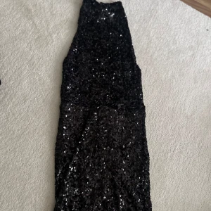 Svart paljettklänning med hög hals - Säljer en svart långklänning täckt av glittrande paljetter. Klänningen har hög hals och är ärmlös, perfekt för att sticka ut på festen. Helt öppen i ryggen.