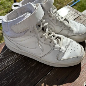 Vita höga sneakers från Nike - Säljer ett par vita höga Nike sneakers med snörning och kardborreband upptill. Skorna har klassisk Nike-logga på sidan och perforerade detaljer på tån för extra ventilation. Perfekta för dig som gillar sportig och stilren look.