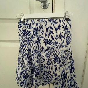 Vit och blå kortkjol - Säljer en söt kortkjol med blått blommigt mönster på vit botten. Kjolen har volanger och resår i midjan för en bekväm passform. Perfekt för varma dagar och enkel att matcha med en enkel topp. Skriv för fler bilder och går att diskutera pris!!
