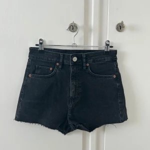 Svarta högmidjade jeansshorts - Svarta jeansshorts med hög midja och råa kanter. Klassisk femficksmodell med knapp och dragkedja framtill. Perfekta för sommaren och enkla att matcha med olika toppar. Aldrig använda. Nyskick! 