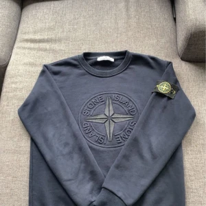 Mörkblå sweatshirt från Stone Island - Säljer en mörkblå sweatshirt från Stone Island med det klassiska broderade kompassmärket på bröstet och avtagbar logotyp på ärmen. Tröjan har rund hals och ribbade muddar vid ärmslut och nederkant. Perfekt för en stilren och avslappnad look. Pris kan diskuteras 