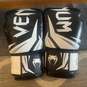 Svarta och vita boxningshandskar från Venum - VENUM CHALLENGER 3.0 BOXNINGSHANDSKAR 14 oz. Säljer ett par snygga boxningshandskar från Venum i svart och vitt med coolt ormmotiv på handleden. Handskarna har bred kardborrestängning och är 14oz. Perfekta för träning och kampsport. 