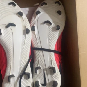 Röda Nike fotbollsskor med svarta detaljer - Säljer ett par röda Nike fotbollsskor med svarta snören och detaljer. Skorna har dobbar för bra grepp på planen och en sportig design med mönstrad ovandel. Perfekta för dig som vill sticka ut på fotbollsplanen!