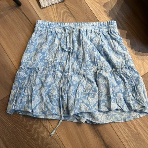 Blå kortkjol med paisleymönster och volang - Säljer en ljusblå kortkjol med vitt paisleymönster. Kjolen har resår i midjan med knytband och volangdetalj runt om. Perfekt för varma dagar och enkel att styla med en enkel topp. Har även shorts under. 