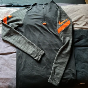 Svart träningströja och byxa från Nike med orange detaljer - Säljer en svart långärmad träningströja och byxa från Nike med halv dragkedja och orange logga på bröstet och ärmen. Ärmarna har en diskret mönstring i grått och tröjan är perfekt för sport och träning.