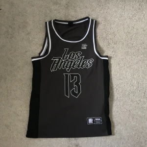 Svart basketlinne Los Angeles FSBN XS - Snyggt svart basketlinne från FSBN i storlek XS med vit text 'Los Angeles' och siffran 13 framtill. Kontrasterande vita kanter runt hals och ärmhål. Perfekt för sportiga vibes och streetstyle. Tillverkat i polyester för en luftig känsla.