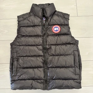 Svart dunväst från Canada Goose - Snygg svart dunväst från Canada Goose med klassisk logga på bröstet. Västen har hög krage, horisontella sömmar och dragkedja framtill. Perfekt att slänga över en hoodie för extra värme. Materialet är glansigt och lätt, vilket gör den smidig att bära.