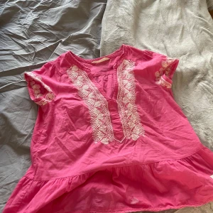 Rosa blus med broderade hjärtan - Supersöt rosa blus med vita broderade hjärtan och mönster längs v-ringningen och ärmarna. Blusen har korta ärmar och volang nertill, perfekt för en färgglad look. Luftig och skön modell som sticker ut med sina detaljer.