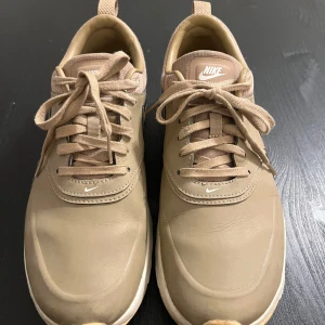 Beige Nike Air Max Thea sneakers storlek 39 - Snygga beige Nike sneakers i skinn med klassisk swoosh på sidan och perforerad häl. Skorna har snörning, vit sula och diskreta detaljer i ljusrosa vid hälen. Perfekta för en clean och stilren look.