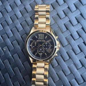 Guldig Michael Kors klocka - Snygg klocka från Michael Kors i guldigt rostfritt stål med svart urtavla och romerska siffror. Klockan har flera små urtavlor och en robust länkarmband som ger en lyxig känsla. Perfekt accessoar för dig som gillar stilrena detaljer.