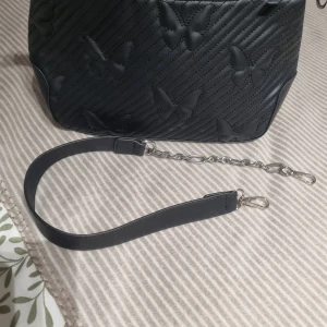 Svart axelväska med fjärilsmönster - Snygg svart axelväska med quiltat fjärilsmönster och diagonala sömmar. Väskan har en avtagbar axelrem i skinn och silverfärgade kedjedetaljer. Perfekt för dig som vill ha en unik och stilren accessoar.