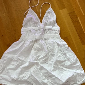 Vit kort sommarklänning med broderi - Superfin vit kort klänning med tunna axelband och broderade blommor. Klänningen har v-ringning och resår i midjan för en snygg passform. Perfekt för varma dagar och har en luftig känsla.