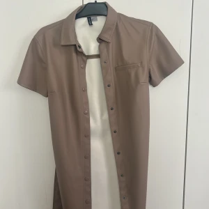 Beige skjortklänning i skinnimitation H&M - Snygg beige skjortklänning från H&M Divided i skinnimitation. Kortärmad modell med knappar hela vägen framtill och tillhörande bälte i midjan. Perfekt för dig som gillar en clean och modern look. Aldrig använd 