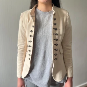 Beige kavaj med dekorativa knappar - Snygg beige kavaj med coola dekorativa knappar i två rader framtill. Jackan har figurnära passform, markerade axlar och lång ärm. Perfekt för dig som vill ha en stilren men ändå unik look.